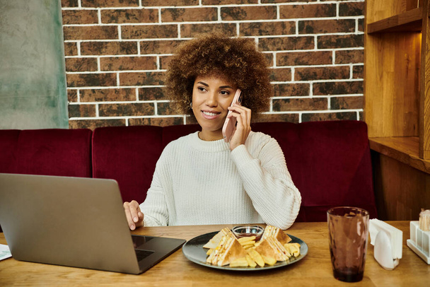 Μια Αφρο-Αμερικανίδα κάθεται σε ένα τραπέζι με ένα λάπτοπ και ένα πιάτο φαγητό, επικεντρωμένη στη δουλειά της.. - Φωτογραφία, εικόνα