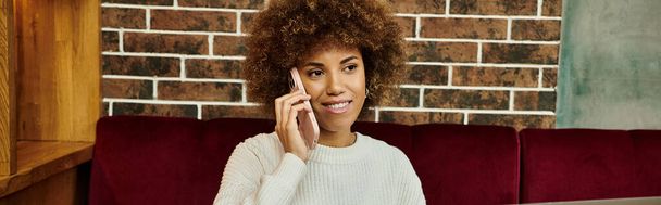 Een stijlvolle Afro-Amerikaanse vrouw zit op een moderne cafe bank, bezig met een telefoongesprek. - Foto, afbeelding