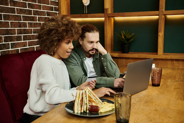 Ein Mann und eine afroamerikanische Frau sitzen an einem Tisch, vertieft in den Inhalt eines Laptop-Bildschirms in einem trendigen Café. - Foto, Bild