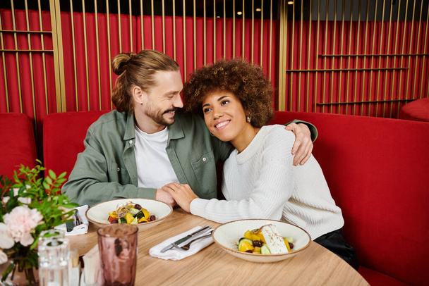 Афро-американская женщина и мужчина наслаждаются обедом вместе за столом, наполненным вкусной едой в современном кафе. - Фото, изображение