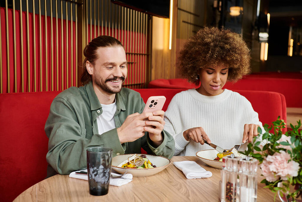 Ένας άνδρας και μια γυναίκα, απορροφημένοι με ένα κινητό τηλέφωνο και φαγητό σε ένα τραπέζι καφέ. - Φωτογραφία, εικόνα