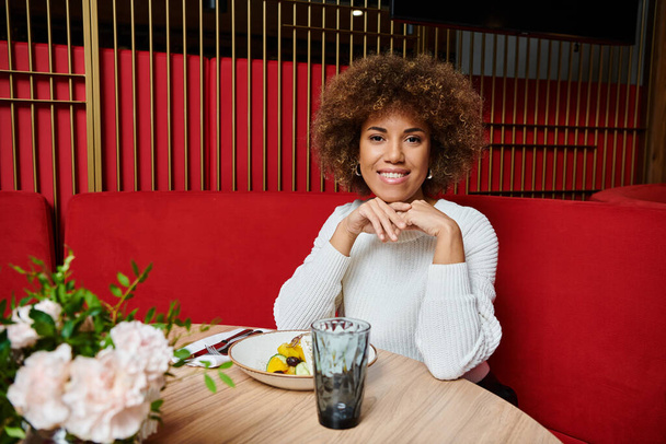 Una donna afroamericana siede a un moderno tavolo da caffè, gustando con grazia un piatto di cibo delizioso.. - Foto, immagini