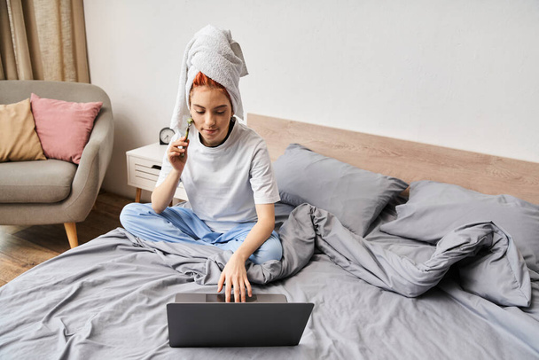 séduisante personne queer aux cheveux rouges en vêtements de maison en utilisant un rouleau facial tout en se relaxant sur le lit avec un ordinateur portable - Photo, image