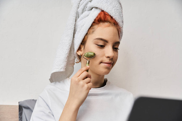 attractive red haired queer person in homewear using face roller while relaxing on bed with laptop - Photo, Image