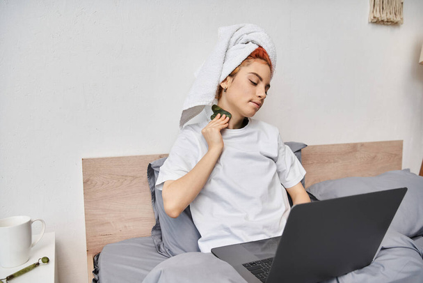 fröhliche ziemlich queere Person mit Haartuch Filme auf Laptop und mit gua sha im Bett - Foto, Bild