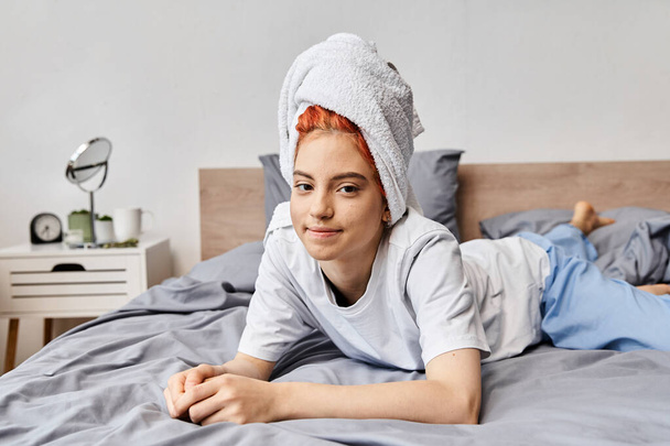 radosna, piękna, queer osoba z ręcznikiem do włosów chłodzi się w łóżku i uśmiecha się do kamery, rano - Zdjęcie, obraz