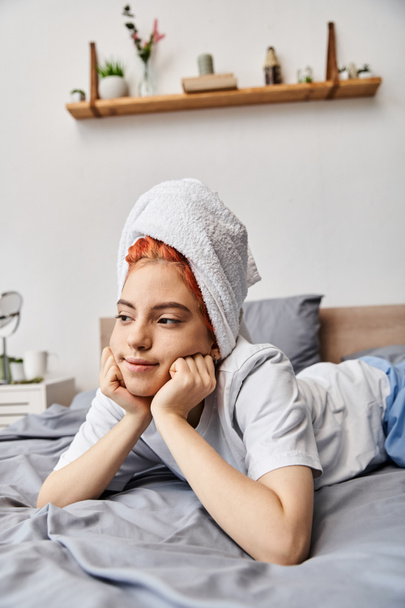 веселая красивая педик в домашней одежде с полотенцем для волос охлаждения в постели и глядя в сторону - Фото, изображение