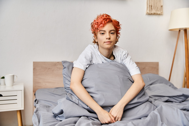 sonolento extravagante queer pessoa em homewear com cabelo vermelho sentado na cama e olhando para a câmera - Foto, Imagem