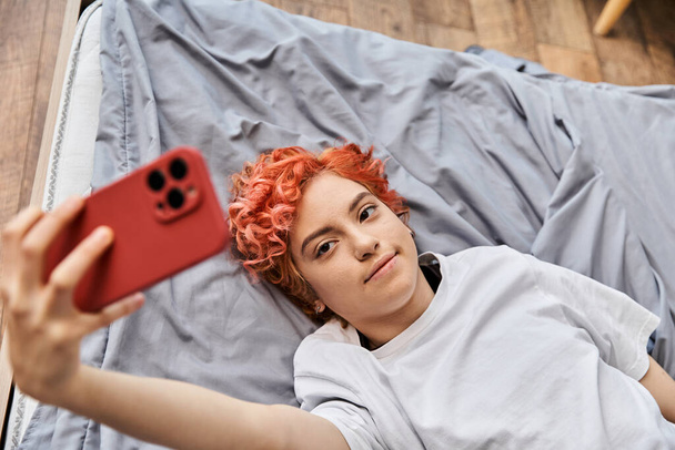 ベッドの上に横たわり,彼女の携帯電話でセルフィーを取っている赤い髪の自家製の若いリラックスしたクイーラーの人 - 写真・画像