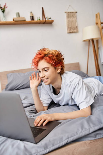 good looking red haired cheerful queer person in homewear having video call while lying in bed - Photo, Image