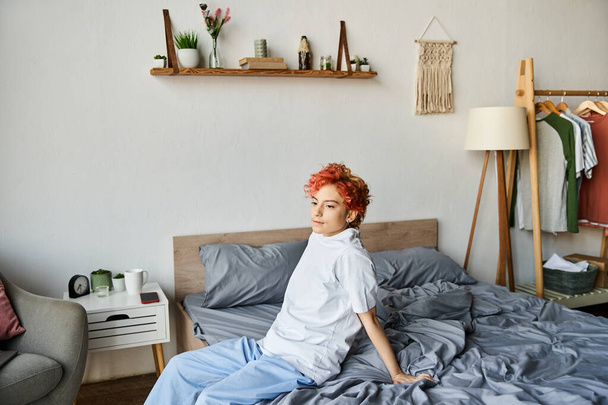 csinos extravagáns személy fehér pólóban vörös hajjal ül az ágyán, szabadidő - Fotó, kép