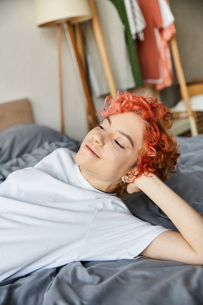 mooie extravagante persoon met rood levendig haar liggend in haar bed en wegkijkend, vrije tijd - Foto, afbeelding