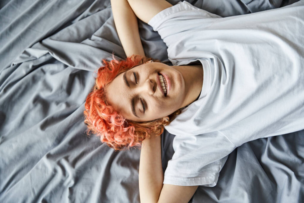 goed uitziende vrolijke homo in gezellige huiskleding ontspannen op haar bed met gesloten ogen, vrije tijd - Foto, afbeelding