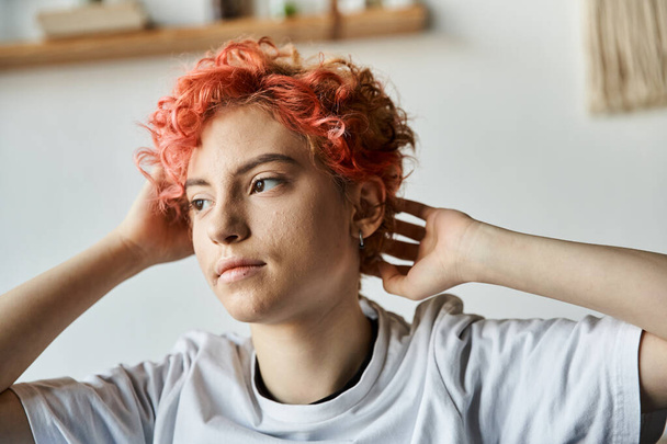 attraktive queere Person mit leuchtend roten Haaren, die auf dem Bett sitzt und wegschaut, Freizeit - Foto, Bild