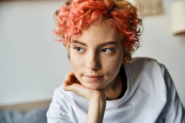 attraente persona queer con i capelli rossi vibranti seduti sul letto e guardando altrove, tempo libero - Foto, immagini