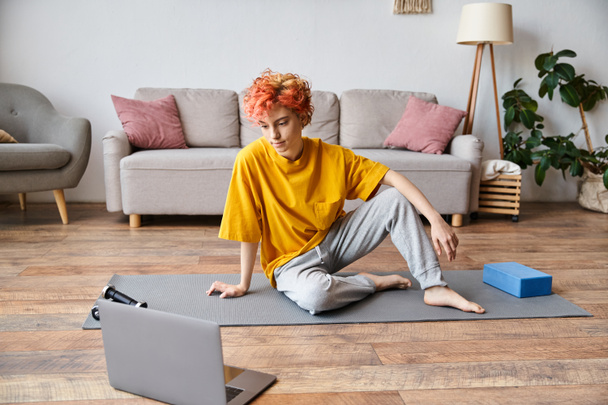 schöne queere Person in gelbem T-Shirt, die auf Yogamatte sitzt und vor dem Sport auf Laptop schaut - Foto, Bild