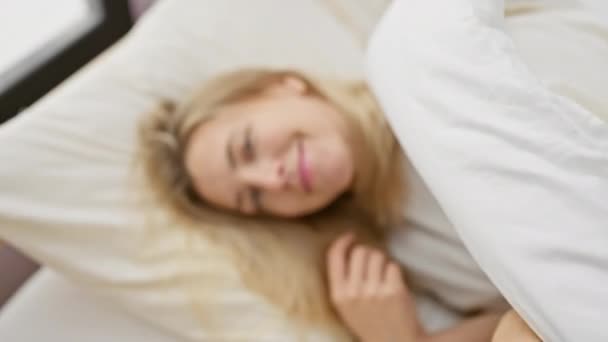 Νεαρή ξανθιά γυναίκα χαμογελά στο κρεβάτι, που απεικονίζει την άνεση και τη χαλάρωση σε ένα φωτεινό εσωτερικό χώρο υπνοδωμάτιο. - Πλάνα, βίντεο