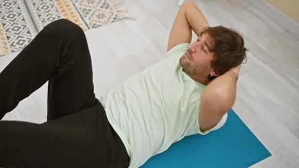 Un joven caucásico se ejercita sobre una alfombra azul dentro de una habitación luminosa, combinando fitness, salud y un estilo de vida casual. - Metraje, vídeo