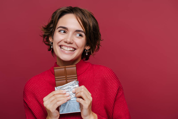 Młoda szczęśliwa kobieta w czerwonym swetrze trzymająca czekoladę i odwracając wzrok na czerwonym tle studio portret. Koncepcja żywienia ludzi  - Zdjęcie, obraz