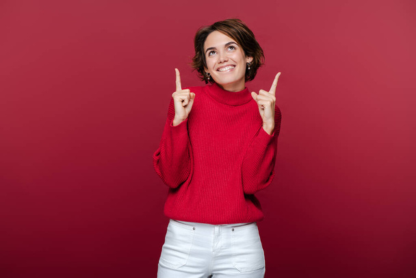 Portret van een positieve jonge vrouw in casual outfit met wijzende vingers omhoog, kopieer ruimte geïsoleerd op rode achtergrond. Winkelen, verkoop, zwarte vrijdag concept  - Foto, afbeelding