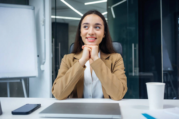 Νεαρή επιχειρηματίας που ξαναζεί ευτυχισμένες αναμνήσεις κατά τη διάρκεια ενός διαλείμματος καφέ κάθεται στο γραφείο της στο γραφείο κοιτάζοντας στο πλάι με ένα χαμόγελο ικανοποίησης και ευχαρίστησης - Φωτογραφία, εικόνα