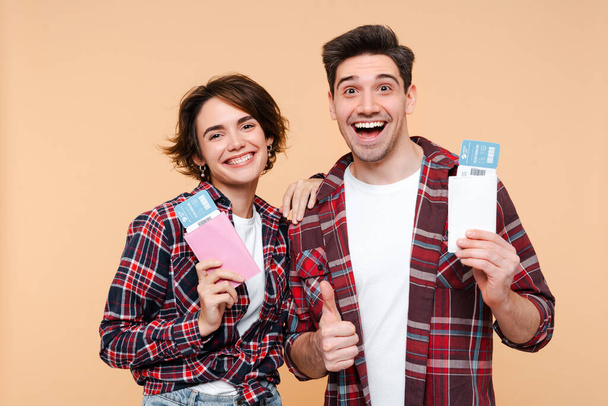 Cheerful couple passenger tourists going for weekend getaway, smiling at camera, posing over beige background with a passports and airplane tickets, awaiting to board the flight  - Photo, Image