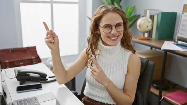 Elegante jonge vrouw glimlacht vrolijk naar de camera terwijl ze naar de zijkant wijst met beide handen op kantoor - Video