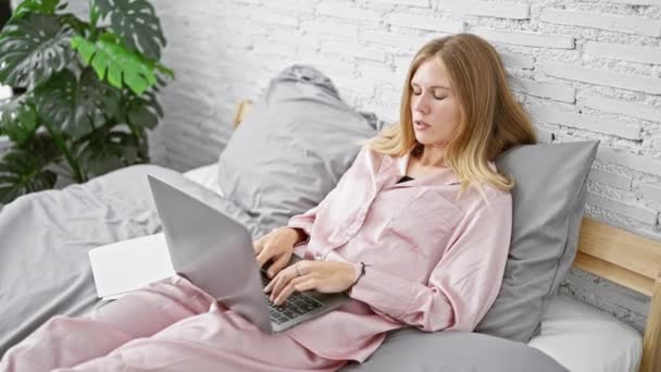 Blondi nainen vaaleanpunainen pyjama työskentelee kannettavan samalla makuuasennossa sängyssä valkoinen tiili tausta. - Materiaali, video