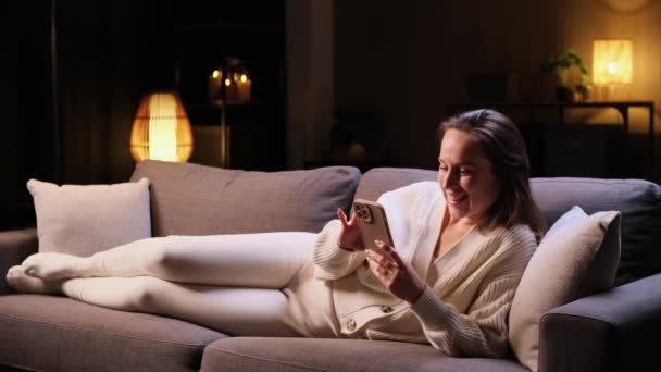 Mujer positiva caucásica acostada en el sofá con teléfono en casa por la noche. Pasar tiempo libre, entretenimiento en línea, navegar y navegar por Internet. - Imágenes, Vídeo