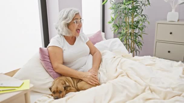 Harmaahiuksinen, keski-ikäinen nainen, juhlii iloisesti voittoaan innoissaan koiran kanssa, istuu sängyllä, yllään pyjama, makuuhuoneessa. Hänen voittoisa hymynsä ja ilmeensä huutaa "voittaja" onnella. - Materiaali, video