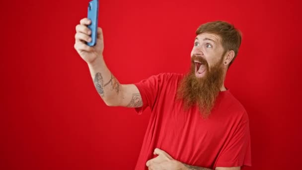 Vreugdevolle jonge roodharige man, vol vertrouwen glimlachend over een toevallige oproep op zijn gadget, geïsoleerd staand tegen een levendige rode achtergrond. geluk straalt als hij geniet van online conferentie. - Video