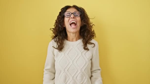 Sprankelende Spaanse vrouw van middelbare leeftijd in een bril, ziend van frustratie, schreeuwend van woede. woedend en bedroefd weergalmt haar agressieve geschreeuw tegen een grimmig, geel geïsoleerd decor. - Video
