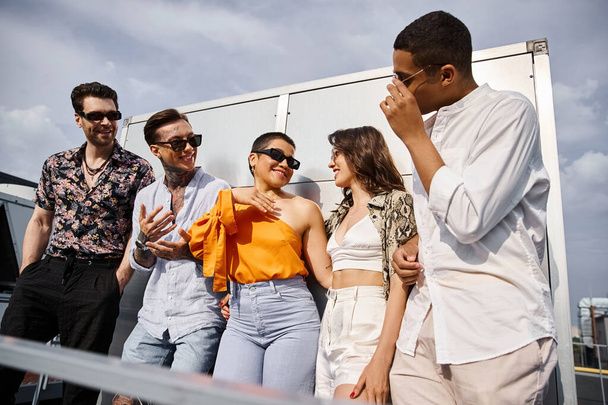 cheerful diverse friends in urban attires with sunglasses posing on roof and smiling at each other - Foto, imagen