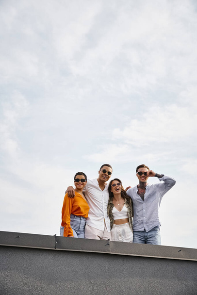four diverse jolly friends with sunglasses in vivid attires posing on rooftop and smiling at camera - Фото, изображение