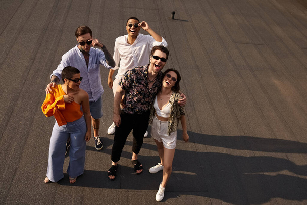 cinq amis gais interracial en vêtements vibrants posant sur le toit et souriant joyeusement à la caméra - Photo, image