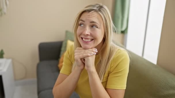 Fröhliche junge blonde Frau im T-Shirt, zu Hause verschämt kichernd, die Hände den Mund verdeckend, seitwärts blickend mit einem funkelnden Auge. - Filmmaterial, Video