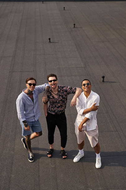 three diverse cheerful male friends with sunglasses posing on rooftop and looking at camera - Foto, Imagem