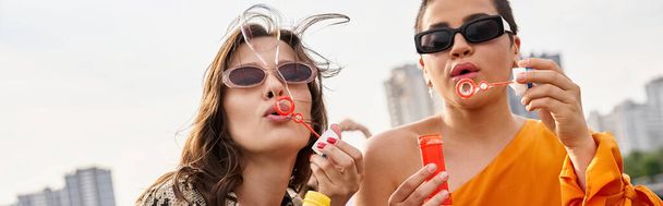 beautiful jolly women in vibrant outfits with sunglasses blowing soap bubbles on rooftop, banner - Photo, Image