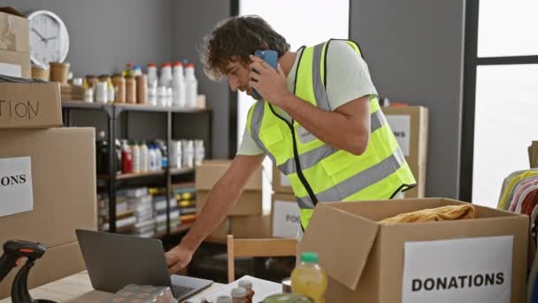 Een man met een goed zichtbaar vest is aan het multitasken op een telefoon en laptop temidden van dozen met het label 'donatie' in een georganiseerde opslagruimte. - Video