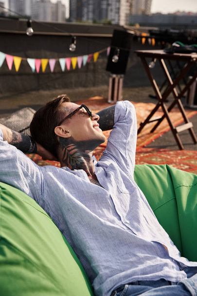 atractivo hombre alegre en traje casual con gafas de sol y tatuajes relajante en el techo y mirando hacia otro lado - Foto, Imagen