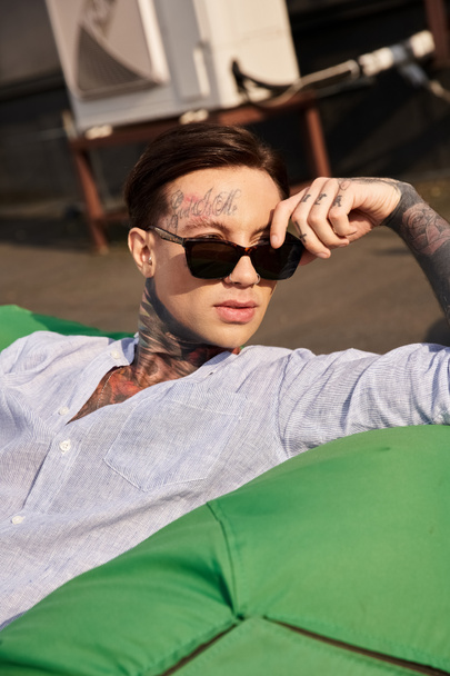 привлекательный молодой человек в повседневной одежде с татуировками и стильными солнцезащитными очками, позирующий на крыше - Фото, изображение