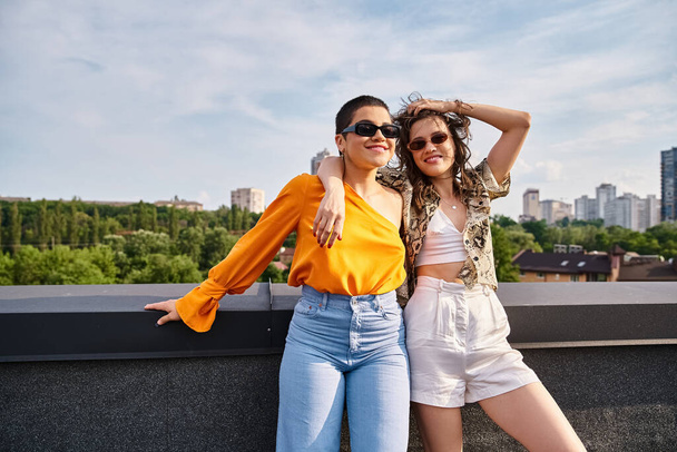 atrakcyjne wesołe kobiety w żywych ubraniach ze stylowymi okularami przeciwsłonecznymi uśmiechnięte do kamery na dachu - Zdjęcie, obraz