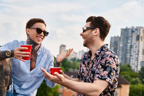 two cheerful men in casual attire with stylish sunglasses talking and holding red cups with drinks - Foto, Imagem