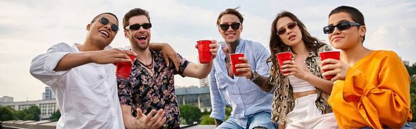 ποικίλες χαρούμενες φίλες με γυαλιά ηλίου κρατώντας κόκκινα φλιτζάνια και χαμογελώντας στην κάμερα στην ταράτσα, πανό - Φωτογραφία, εικόνα
