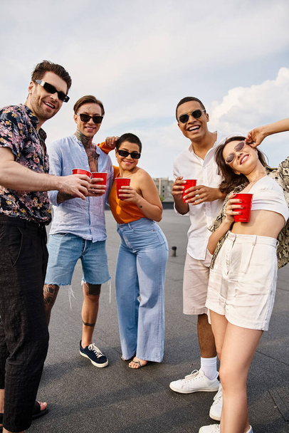 joyful multicultural friends in vibrant clothes holding red cups with drinks and smiling at camera - Фото, изображение