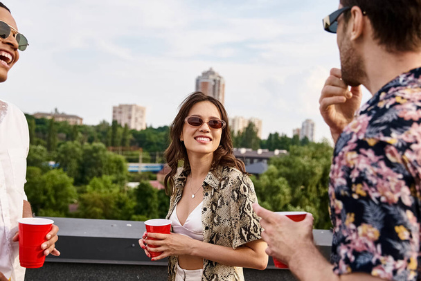 vreugdevolle interraciale vrienden in casual kleding met een zonnebril drinken uit rode kopjes op het dak - Foto, afbeelding