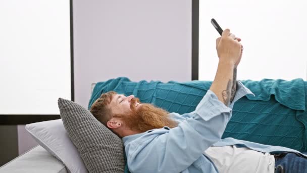 Vidám, fiatal vörös hajú férfi boldogan gépel üzeneteket az okostelefonján, kényelmesen fekszik a kanapén, elveszve az internet vidám világában, közvetlenül otthonos nappalijának szívében. - Felvétel, videó