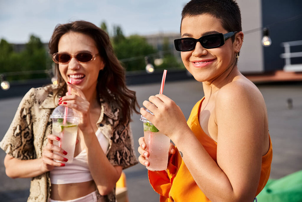 cheerful beautiful female friends drinking cocktails at rooftop party and smiling at camera - Photo, Image