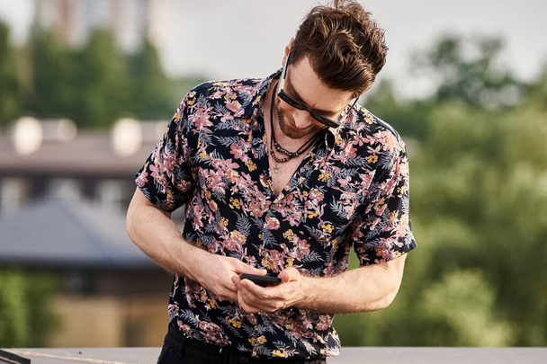 vreugdevolle knappe man met een zonnebril in levendige kleding kijkend naar zijn telefoon op het dak - Foto, afbeelding