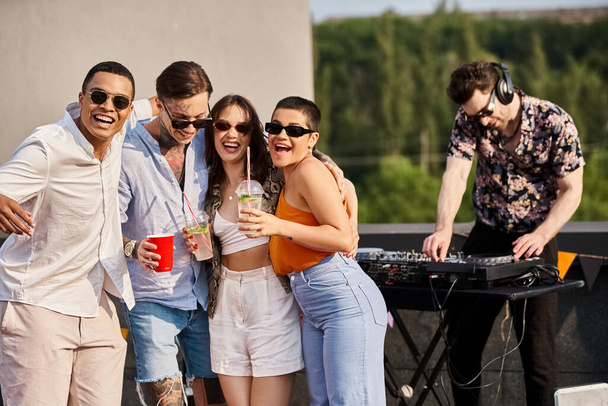cheerful interracial friends with drinks posing next to DJ at rooftop party and smiling at camera - Foto, Imagen